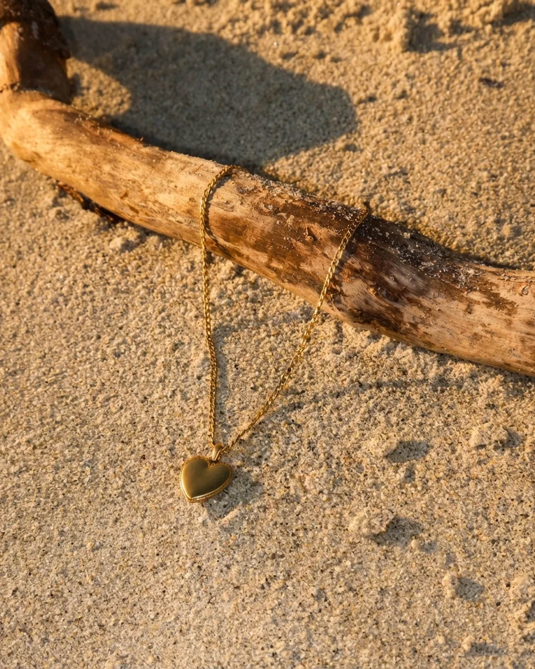 A heart pendant necklace draped over driftwood on sand
